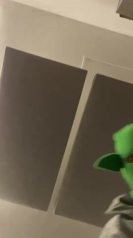 A person wearing a bright green balaclava with elf-like ears is standing in a room. The room appears to be a residential space, possibly a bedroom, with a white or light-colored ceiling, gray or white walls and furniture. The video appears shaky and focuses on the masked person. There is a brief glimpse of a bed with white bedding. The individual's actions suggest they may be dancing or moving to music, though the audio is distorted and indistinct.