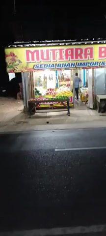 A fruit stand named "MUTIARA BUAH" is visible at night. The sign indicates it sells "SEDIA BUAH IMPOR & LOKAL," meaning imported and local fruits. The stand is illuminated, displaying a variety of fruits, including apples, watermelons, and other produce. A person, wearing a head covering, is standing inside the fruit stand. The stand is situated alongside a dark, wet road, suggesting it may have recently rained. There are also some wooden tables and a trash can visible near the stand.