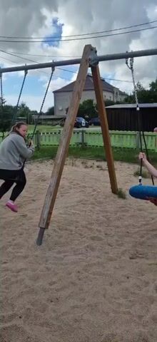 A video taken at a playground shows two children on swings. The playground has a sand-covered ground and a wooden A-frame swing set with metal poles. A girl with long brown hair, wearing a grey hoodie and black leggings, is on the left swing, moving back and forth. A boy with blonde hair, wearing a white t-shirt, is on the right swing, which is a nest-style swing. He is facing the camera and holding the ropes. A younger child wearing a pink hoodie and hat is standing in the foreground, facing the boy on the swing, appearing to observe or interact. In the background, there are green fences, trees, and buildings. The sky is cloudy. The video spans approximately five seconds.