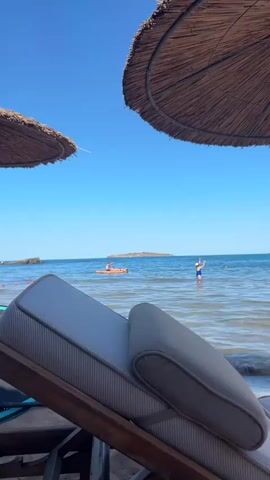 The video shows a peaceful beach scene on a clear, sunny day. The perspective is from a lounge chair, looking out towards the ocean. Two straw umbrellas are visible in the foreground, providing shade.

In the water, several people are enjoying the day. One person is standing waist-deep in the water, wearing blue swim trunks and a white hat. Further out, two people are on an orange paddleboard. One person is sitting and appears to be paddling with a long oar, while another person is lying down on the board. In the distance, a small island with some structures on it can be seen.

The water is calm and blue, with gentle waves. The sky is a vibrant, cloudless blue. The overall atmosphere is one of relaxation and leisure. The bright sunlight and clear sky suggest it is daytime, likely mid-morning or afternoon. The presence of lounge chairs and umbrellas indicates a resort or public beach area. The scene evokes a sense of a pleasant summer vacation. There is no visible text in the media file.