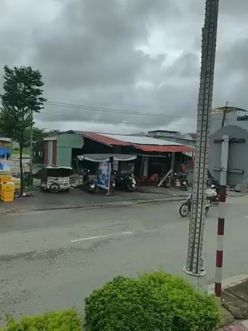 A cloudy day scene captures a street with several buildings and shops. A motorcycle carrying two people is seen riding on the road. In the background, a hotel with a sign that reads "PHUONG HOANG HOTEL" is visible. A road marker with mileage information for Dak To and Ho Chi Minh City is also present. Several parked motorcycles, market stalls with umbrellas, and some greenery are part of the roadside environment. Trucks and vans are also present on the road. A circular traffic sign on a pole stands near the roadside.