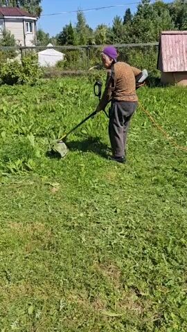 An elderly woman with white hair, wearing a purple headscarf, brown sweater, and dark pants, uses a string trimmer to cut grass in a sunny, outdoor setting. She is standing in a yard with green grass and some overgrown weeds. In the background, there is a fence, a building, and trees under a blue sky. The woman is holding the trimmer with both hands and is actively engaged in the task.
