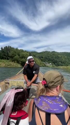 A group of people are on a boat traveling down a river. The sky is blue with white clouds. The riverbanks are lined with trees and hills. A man in the front of the boat is rowing. Two people are sitting behind him, facing forward. The man is wearing a black t-shirt, gray shorts, and a white hat. The people behind him are wearing casual clothing and hats. The boat is wooden and appears to be a traditional Japanese river boat. There are Japanese characters on a sign on the side of the boat.