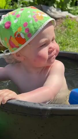 A baby with blonde hair and a green and pink sun hat is sitting in a black tub full of water. The baby is shirtless and is holding a blue toy watering can in its right hand. The baby looks around with a happy expression and is splashing the water. The background is a grassy area with some plants.