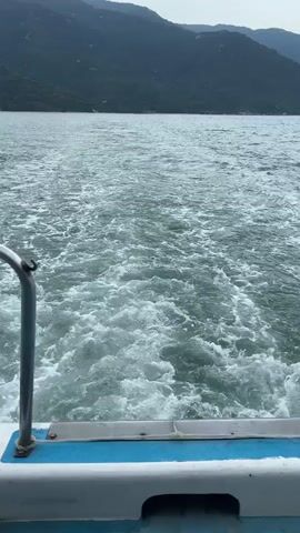 This video shows the wake of a boat moving through water. The camera is positioned at the back of the boat, looking towards the wake and a mountainous coastline in the distance. The water is choppy and foamy due to the boat's movement. The sky is overcast.