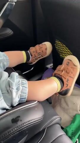 The video captures a close-up view of a toddler's legs and feet in a car. The child is wearing light blue denim overalls and brown sandals with black and yellow striped socks. The child's feet are moving, suggesting they are kicking or playing. The background shows the interior of a car, including a black leather seat, a yellow mesh pocket on the back of a seat, and a purple bag. A green item of clothing is also visible near the child's feet. The overall impression is one of a child being in a car seat, possibly during a journey. The text "00:00" to "00:04" appears at the beginning of the video, indicating timestamps. The context provided suggests the location is Srednyaya Toyma, Russia.