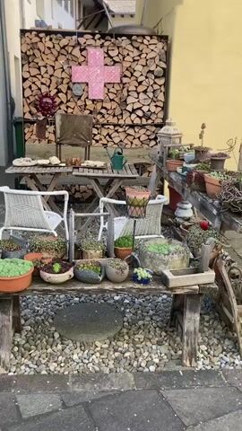 The video shows an outdoor area, possibly an alleyway or courtyard, with a rustic and well-decorated ambiance. The scene features a gravel ground and stone pavers.

On the right, a large stack of firewood forms a wall, against which a bright pink cross is displayed. In front of the firewood, there's a wooden table with two chairs, and a green watering can sits on the table. A green recycling bin is partially visible behind the table.

In the foreground, a long wooden bench is heavily laden with numerous potted plants, predominantly succulents, in various decorative containers. Two flat, round stepping stones are placed on the gravel in front of this bench.

As the camera pans left, a large blue barn-style door with a decorative wreath on it comes into view, set into a white building wall. To the left of the blue door, another wooden plant stand holds more potted plants. No people are visible in the video. The lighting suggests it is daytime.