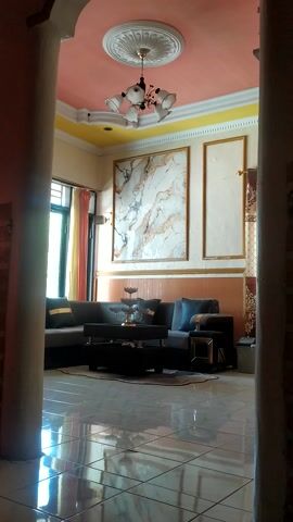 This media file depicts an interior view of what appears to be a living room or lounge area. The camera angle is from a high vantage point, looking down into the room.

**Subjects:**
*   **Objects:** The room is furnished with a dark-colored sofa and a coffee table. There's a chandelier with multiple lights hanging from the ceiling. The walls are decorated with framed artwork, one of which features a marble-like pattern. The floor appears to be tiled. A window with curtains is visible.
*   **Scene/Setting:** The scene is an indoor living space. The walls are painted in pastel colors, with a combination of pink and yellow. The ceiling has decorative molding and a central medallion.

**Activity/Event:**
No discernible activity or event is taking place; the scene appears static, suggesting it might be a still photograph or a paused video frame.

**Notable Details:**
*   **Lighting:** The room is illuminated by a combination of the chandelier and natural light coming from the window. The lighting suggests it could be daytime.
*   **Decor:** The decor appears to be somewhat formal, with the framed artwork and chandelier contributing to this impression. The marble pattern artwork is a prominent visual element.
*   **Color Palette:** The dominant colors are soft pastels (pink, yellow) on the walls, contrasted with the darker furniture and the marble pattern artwork.
*   **Location Cues:** While specific location cues are not directly visible within the frame, the provided context indicates the location is Desa Penggalang Kecamatan Adipala, Indonesia. This detail, however, is external to the visual content of the media itself.

**Visible Text:**
No visible text is present in the media file.