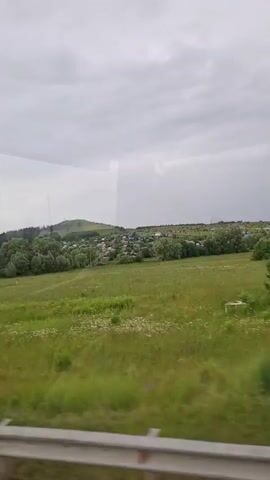 The video captures a view from a moving vehicle, looking out at a rural landscape under a cloudy sky. In the foreground, a metal barrier runs horizontally across the bottom of the frame. The middle ground features a vast, green field dotted with small white wildflowers. Beyond the field, a cluster of trees forms a dense line, and further in the distance, a small village with houses and roofs is visible nestled against a gently rolling hill. The hill is covered in trees, creating a lush, green backdrop. Power lines and a utility pole are also visible in the middle ground. The overall impression is one of natural beauty and tranquility in a rural setting.