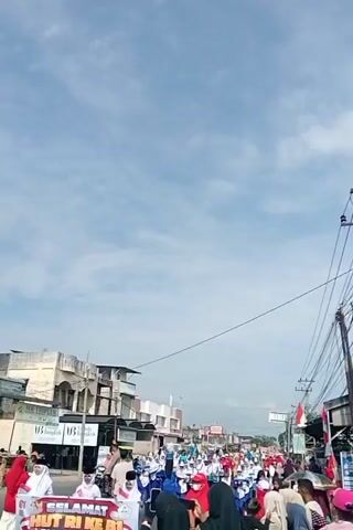 This is a vertical, eye-level, medium shot video of a parade passing by. The parade is on a street lined with buildings and sidewalks. People are gathered on the sidewalks watching the parade. Many people in the parade are wearing white uniforms with blue accents, and some are carrying flags or musical instruments. In the foreground, several people are watching the parade, some holding up cell phones to record. The sky is blue with some clouds. There are electrical wires and utility poles visible along the side of the street. One building on the left has text that reads "SELAMAT ... RI KE B1..."