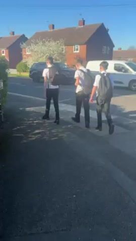 The video shows a residential street corner on a bright, sunny day. Three young males, presumably school students, dressed in black pants, white shirts, and black shoes, are walking across a zebra crossing. They are all wearing backpacks. As they cross the street, another person joins them from behind, and they continue walking. A street sign labeled "Harding Avenue" is visible on the corner. Several cars are parked along the side of the road. Houses with brick facades and some with solar panels line the street. The sky is clear and blue. The camera follows the students as they walk.