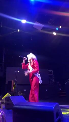 A drag queen in a red dress and a sash that reads "FEMME FATALE" stands on a stage, holding a microphone and singing. Blue and white spotlights shine on the performer, creating a dramatic effect. The stage is dimly lit, with speakers and other equipment visible in the foreground. In the background, a large screen and other stage elements can be seen. The drag queen's wig is blonde and voluminous, and they are wearing a crown. The sash is white with gold lettering. The performer's left hand is raised in a gesture as they sing. The overall atmosphere appears to be that of a performance or competition.