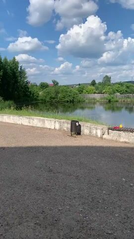 A still shot captures a body of water with a reflection of the sky and trees. The water is surrounded by lush green vegetation and trees, and in the background, there are houses with red roofs and other structures. The sky is blue with fluffy white clouds. In the foreground, there is a concrete structure with a metal grate, and a trash can. The ground in the immediate foreground is covered in dark grey gravel.