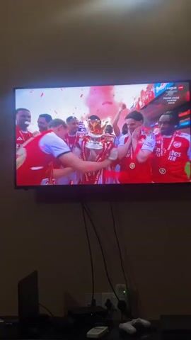 Here is a detailed description of the video:

The video shows a TV screen displaying a live broadcast of Arsenal football players celebrating their Premier League championship win. The players, dressed in their red and white jerseys, are gathered around the trophy, which is prominently displayed in the center. Confetti in red and pink colors fills the air, adding to the celebratory atmosphere. The players can be seen cheering, clapping, and lifting the trophy.

Below the TV, there is a shelf or table with various electronic devices. A laptop is visible on the left side, and there are video game controllers and what appears to be a game console. The room is dimly lit, with the TV being the primary light source. The walls are a neutral color.