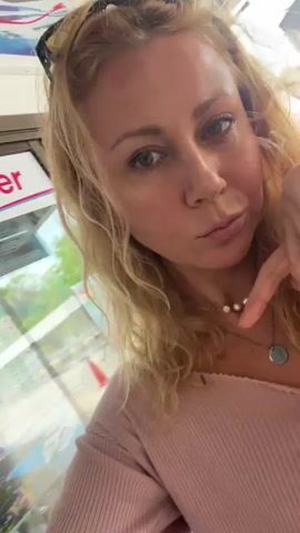 A woman with blond, wavy hair and green eyes stands inside, possibly in a transportation terminal given the "Ticket Counter" sign visible. She wears a pink, ribbed top and a double necklace of pearls and a silver pendant. She touches her hair, which is partially pulled back by a black headband, and adjusts the sleeve of her top. She looks directly at the camera with a small smile. A light brown bag strap is visible over her shoulder.