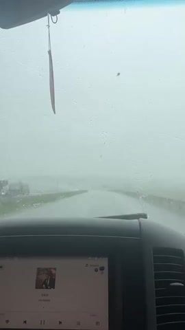 The video is filmed from the interior of a vehicle looking out through the windshield. Heavy rain is falling, evident by the water droplets and streaks on the glass, and the windshield wipers are in motion. The road ahead is wet and appears to curve to the right. Visibility is significantly reduced due to the rain and fog. The dashboard of the vehicle is visible in the foreground, with a screen displaying what appears to be a music application. A dangling air freshener is hanging from the rearview mirror. The overall environment suggests a very inclement weather day.