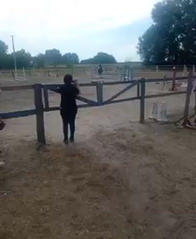 A person holding a camera stands with their back to the viewer, leaning on a wooden fence. Another person stands to the left of the camera holder. In the background, a person on a horse jumps over an obstacle. The scene takes place in a dirt arena with more fences and obstacles visible. The sky is overcast.