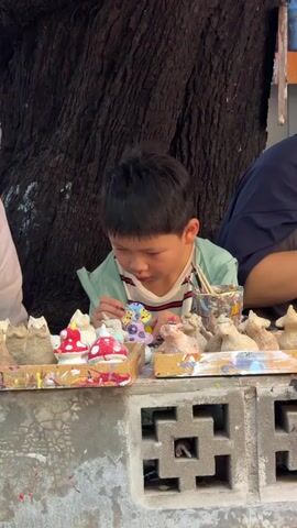 This media file shows a close-up view of a young boy intently painting small, ceramic figures. The boy, with dark hair and a focused expression, is wearing a light green jacket over a striped t-shirt. He is holding a paintbrush and delicately adding color to a small, purple and yellow figure.

In front of him, arranged on a wooden tray, are several unpainted and partially painted ceramic figures. These include mushroom-shaped objects with red caps and white dots, and other small animal-like or whimsical figures. A cup filled with paintbrushes and various colors of paint is visible to his right.

The scene appears to be an outdoor setting, possibly an art workshop or a craft market. Behind the boy is the dark, textured bark of a large tree, suggesting a natural environment. The figures are placed on a textured, light-colored surface that looks like concrete, possibly a table or a low wall.

The lighting suggests it is daytime, and the activity is a creative and engaging one. The boy's concentration and the presence of multiple art supplies indicate he is engrossed in the process of painting. The overall mood is one of quiet creativity and childhood engagement. The location context provided, Ban Sai Kham, Thailand, places this scene within a specific geographical and cultural setting. There is no visible text in the media file.