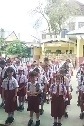 A large group of elementary school children in uniform are standing in a line outdoors. The children are wearing white shirts with red ties, and maroon skirts or shorts. They are all carrying backpacks. In the background, there are buildings that appear to be part of a school campus. Some of the children are looking at the camera, while others are looking forward. The overall scene suggests a school assembly or the start of a school day.