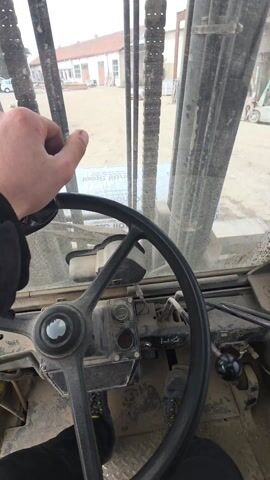 The video shows a view from inside the cab of a large industrial vehicle, likely a forklift or similar machine. The driver's hand is visible on the steering wheel, and the dashboard and controls are also in view. Through the front window, a construction site with a building is visible. The windshield is dusty.