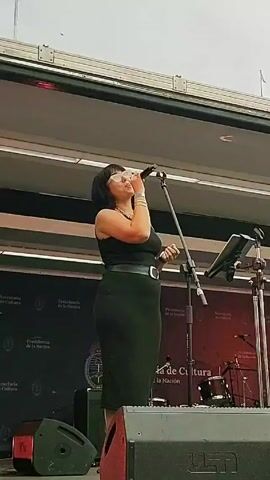 A woman sings on stage, holding a microphone. She wears a black dress and sunglasses. The stage has black panels behind her, with branding, including "Presidencia de la Nacion." There is a microphone stand, a music stand, and a drum kit visible behind her. The lighting is bright, suggesting an outdoor performance during the day.