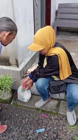 Two women are outside on a stone patio next to a white wall. The first woman, on the left, has gray hair pulled back in a bun and is wearing a white and blue patterned top with a floral skirt. She is wearing red sandals. The second woman, on the right, is sitting on the stone steps. She is wearing a yellow hijab with a cap, a black jacket over a yellow shirt, blue jeans, and gray mesh slip-on shoes. She has a small black purse slung across her body.

A small green plant in a white container sits on the patio. There is a white plastic bag on the ground. The first woman is placing items from the plastic bag onto the stone steps where the second woman is sitting. The second woman is looking down at her purse. There is also some other items, including bread, on the stone step.

Behind the women is a white door and a wooden bench. The ground is covered in small black rocks. A pink and a black plastic object lie on the ground.