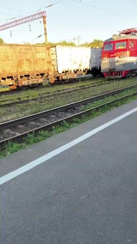 A red and gray electric locomotive is parked on train tracks next to several freight cars. The locomotive is facing the right side of the frame and has its headlights on. A person is visible in the driver's seat of the locomotive. The freight cars are weathered and appear to be made of metal. Overhead power lines are visible above the train tracks. The scene is outdoors under a clear blue sky. The ground in the foreground is a paved surface with a white line marking its edge. Grass and weeds grow between the train tracks.