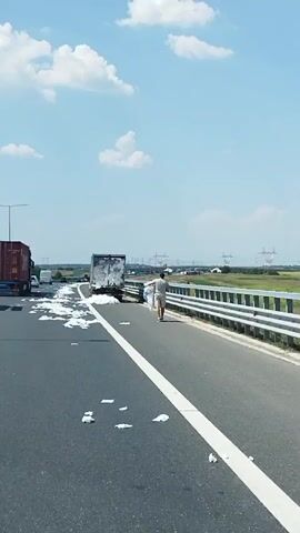 A man is walking along the shoulder of a highway. He is carrying a large, white bundle of something. The highway is mostly empty except for a few other vehicles. White pieces of paper are scattered across the road and shoulder. A truck is parked on the side of the road with its back doors open, and it appears to have been carrying the white material that is now on the road. The sky is blue with some clouds.