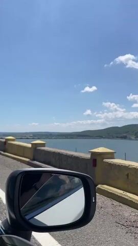A side angle shot from a car shows a body of water with mountains in the background, a sunny sky with some clouds, and a yellow and gray concrete barrier to the left.