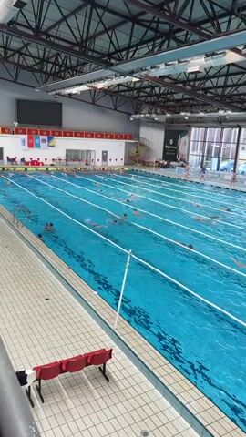 The video shows an indoor swimming pool with multiple lanes. Many people, primarily children, are swimming in the pool, spread across the different lanes. The pool is filled with blue water, and lane markers are visible separating the lanes. Some people are standing or sitting at the edge of the pool. A large window is seen in the background, providing natural light. The building has a high, complex metal roof structure. There are red benches near the edge of the pool on the left side of the frame.