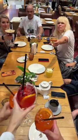 The video shows a group of people sitting around a wooden table in what appears to be a restaurant. They are raising glasses for a toast. In the foreground, three hands are holding glasses with red and orange drinks, garnished with orange slices and straws. In the background, several other individuals are visible. One woman on the left is holding a martini glass. A man in a white t-shirt is seated at the table, and a woman with blonde hair on the right is holding a white wine glass. Another person on the far right is partially visible. The table is set with plates, napkins, a teapot, and cell phones. The lighting suggests an indoor environment, possibly during daytime given the ambient light from windows or overhead fixtures. The overall atmosphere is social and celebratory.