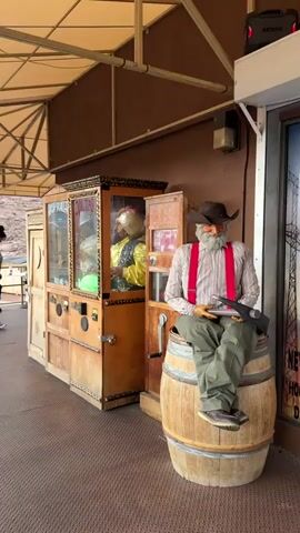 This media file captures an outdoor scene with several subjects and objects. In the foreground, a mannequin dressed as a cowboy sits on a wooden barrel. He wears a cowboy hat, a plaid shirt, green pants, red suspenders, and a brown vest. He is holding what appears to be a pickaxe or shovel.

Behind the cowboy mannequin, there are two vintage-looking arcade machines. One is taller and appears to be a Zoltan fortune teller machine with a glass front displaying figures and text. The other is a smaller machine next to it. These machines are housed under a tan awning, suggesting an outdoor covered area.

In the background, several people are visible. One person is standing and looking towards the arcade machines, while another is seated on a bench. Further in the distance, two people are walking away from the camera. The setting appears to be a desert landscape with rocky hills and sparse vegetation, consistent with the location context of Boulder City, USA. The overall atmosphere seems casual and perhaps tourist-oriented, with the arcade machines and mannequins contributing to a themed environment. The lighting suggests it is daytime, likely late morning or afternoon, with a clear sky. There is no strong indication of weather conditions beyond it being dry.

Visible text includes "HIGH SCORE" and "ZOLTAN" on the fortune teller machine. The overall impression is a quirky roadside attraction or a themed area in a visitor center.