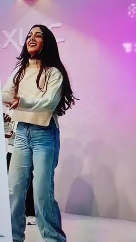A video shows a woman with long dark hair and a beige sweater and jeans dancing. Behind her, a man with dark hair and a light-colored shirt and dark pants is also dancing. The background is a light-colored wall with the letters "XINC" in large white font. The lighting is pink and purple.