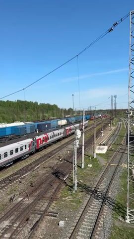 The video presents a high-angle view of a train yard under a clear blue sky. Several tracks converge and run parallel, with trains parked on some. On one track, a passenger train with white and gray cars and the red "РЖД" logo is visible. Adjacent to it are rows of blue shipping containers. The ground is covered with gravel and sparse green vegetation. A tall, lattice-style metal tower stands in the foreground on the right side of the frame. Overhead, power lines stretch across the scene. In the background, a line of trees borders the yard.