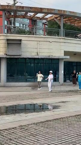 Two people, one wearing a white hat and white athletic clothing and the other wearing a light-colored t-shirt and gray athletic pants, are walking and performing hand movements in unison on a paved walkway in front of a building with large windows. They are both wearing white gloves. The environment appears to be outdoors during the day, with a reflection of trees and other people visible in the windows of the building. The ground in the foreground has a puddle of water.