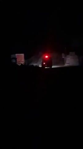 The video captures a nighttime scene with a vehicle in the distance, its red taillight illuminated. The vehicle appears to be on a dirt or gravel surface, with piles of what look like debris or construction materials scattered around. There are hints of structures or walls in the background, also illuminated by red light. The overall environment is dark and appears to be an outdoor, possibly industrial or construction site.