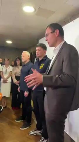 A group of five people is standing indoors. One man is speaking, dressed in a gray suit, white shirt, and glasses. He is gesturing with his hands. Behind him are three people, a woman and two men. The woman wears a light-colored blouse, black pants, and glasses. The two men wear dark-colored shirts and pants. The man furthest back from the speaker appears to have white hair. The other man wears a navy-blue polo shirt with a logo on it. The fifth person, a woman, is standing to the left of the group, wearing a white top and skirt. The walls behind them are painted white. The speaker stands on a wooden floor. The lighting is bright.