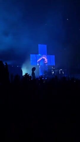 A concert crowd with raised hands is visible in silhouette. A stage with a DJ and various electronic equipment is lit by bright blue and green spotlights, creating beams of light and haze. A cutout figure resembling an alien stands on the left side of the stage. A large, bright blue screen is on the right side of the stage. A disco ball is visible at the top of the frame.