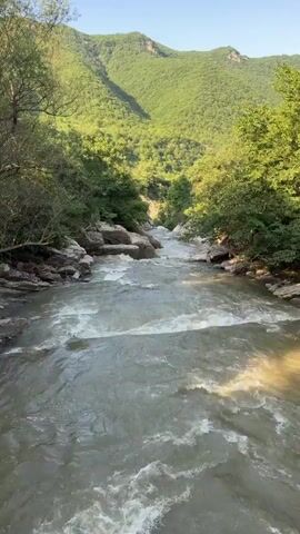 A drone shot shows a turbulent river flowing through a lush green valley. Trees line the riverbanks, and mountains rise in the background. The water is a murky gray-green, with white foam indicating rapids. The sky is clear and blue.