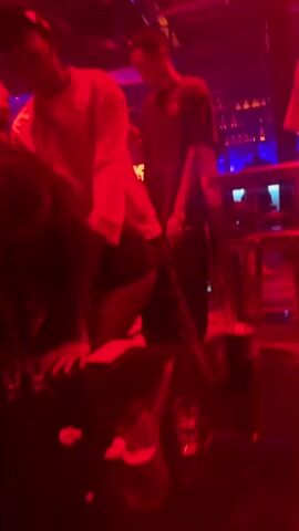 This is a short, shaky video clip filmed in a dimly lit nightclub. The dominant color is red, cast by the club's lighting, with some yellow light highlighting a person in the foreground. This person, wearing a yellow sweatshirt and a baseball cap, appears to be dancing or moving rhythmically. Other figures are visible in the background, blurred by the low light and movement, suggesting a crowded environment. A bar area with bottles is dimly visible in the far background. The audio consists of muffled music and indistinct chatter.