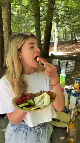 A woman with long blonde hair and a white t-shirt sits outdoors in a wooded area, holding a paper plate with food. She is eating from the plate with her fingers. The plate contains meat, grilled vegetables, and what appears to be a flatbread or cheese. There are other plates, drinks, and bottles on a table next to her. She looks at the camera and smiles, then takes a sip from a clear glass bottle with a yellow liquid.