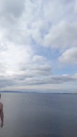 A video shows a wide shot of a body of water under a cloudy sky. There is a dark line of land visible in the distance. In the far right of the frame, a yellow crane is visible. A person's head and shoulders are visible in the lower left of the frame as they look towards the water and sky.