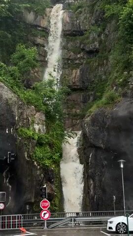 A powerful, muddy waterfall cascades down a steep, rocky cliff face covered in lush green vegetation. The water crashes into a pool at the bottom, creating a significant amount of spray. In the foreground, a traffic light shows a red stop signal, and a "no entry" sign is visible. A white car is parked on the right side of the frame, and a metal railing separates the road from the waterfall area. The scene suggests a recent heavy rainfall that has intensified the flow of the waterfall and made the water appear murky.