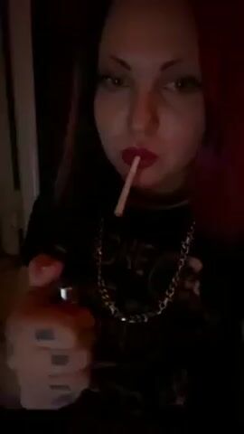 A woman with dark eye makeup and red lipstick lights a thin cigarette or joint. She uses a lighter and takes a drag, exhaling the smoke. She has red hair and wears a black t-shirt with a chain necklace. Her hand has tattoos on it. She looks directly at the camera. The lighting is dim and there is music playing in the background.