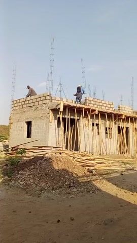 The video shows a single-story building under construction. The walls are made of gray cinder blocks, and workers are on the roof laying more blocks. Wooden beams support the roof's structure. Rebar sticks up from the walls, presumably for reinforcement. A pile of dirt and wood planks lies in the foreground. The ground is mostly dirt, and a clear sky is visible.