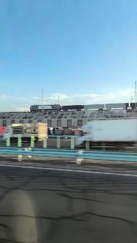 The video shows a passing view from a vehicle traveling along a highway. The highway has multiple lanes separated by guardrails. In the background, a train is visible on an elevated track. Below the train tracks, there are various freight containers and trucks. The sky is clear and blue. The time displayed is between 00:00 and 00:04.