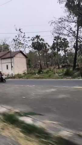 A slightly blurry video captures a roadside scene with sparse vegetation and a few small buildings. A road runs through the center of the frame, with dry grass and dirt on either side. In the background, several trees with bare branches are visible, suggesting a cooler season or a specific type of tree. One of the buildings appears to be a simple, light-colored structure with a pitched roof. Another building, further down the road, has a more open facade with some signage. A blue and white traffic sign, indicating a U-turn is permitted, is positioned on a pole near the road. The lighting is diffused, suggesting an overcast sky or early morning/late evening. There are no people or vehicles visible in the video. The overall impression is a rural or semi-rural setting.