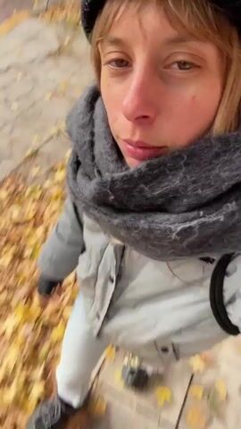 A person wearing a black knit cap, a gray scarf, a shiny gray down jacket, light blue jeans with a black studded belt, and black shoes walks on a sidewalk. The ground is covered in fallen yellow leaves. The person is holding the camera and filming themselves as they walk, showing their face, clothes, and the ground. Keys hanging from a carabiner can be seen attached to the belt.