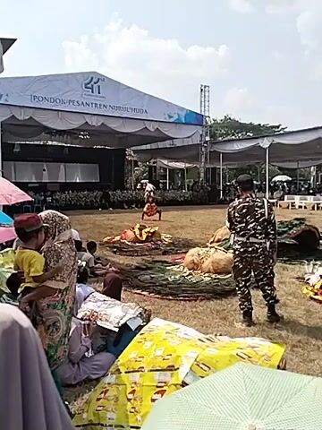 The video shows an outdoor performance taking place on a grassy field. In the background, there is a large stage covered by a white tent, with speakers and lighting equipment visible. A banner with writing in Indonesian is displayed on the tent. A performer wearing a colorful costume and an elaborate headdress with white spikes is dancing in the center of the field. Several other performers, seemingly dressed in similar costumes or props, are lying on the ground in front of the dancing performer. A person in a camouflage uniform is standing to the right, observing the performance. In the foreground, several people are seated on the ground, some of whom are using umbrellas for shade. The overall atmosphere appears to be that of a public event or festival.