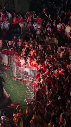 A large crowd of people, mostly young adults, are packed together outdoors. Many are wearing red and yellow clothing, some with Spanish flags, suggesting a celebration or sporting event. They are holding up small Spanish flags and some are waving larger flags. In the middle of the crowd, there are metal barriers separating sections of people. Some individuals are leaning over the barriers. The lighting is dim, indicating it is nighttime or indoors with low light. Some people appear to be cheering or excited, with hands raised in the air. There is a green area visible behind the barriers, possibly grass. The overall atmosphere is energetic and celebratory.